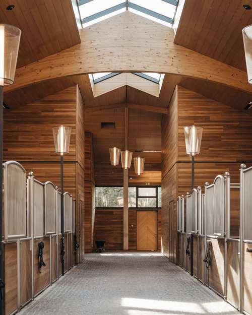 Holzstall mit Pferdeboxen, beleuchtet von großen, modernen Lampen, in einem geräumigen Inneren mit gewölbter Decke und Oberlichtern, die natürliches Licht einlassen. Steinboden unter den Boxen.