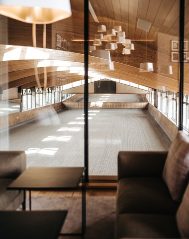 Ein moderner Viebrock Reithallen Innenraum mit hohen, holzgetäfelten Decken und großen Fenstern. Im Vordergrund ist eine gemütliche Sitzecke mit einem Tisch, während im Hintergrund der lichtdurchflutete Reitbereich sichtbar ist.