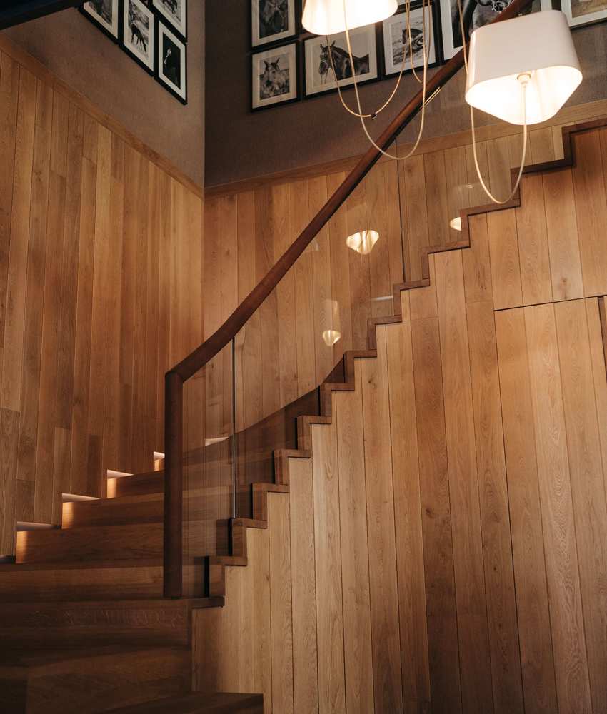Moderne Treppe aus Holz mit Glasgeländer in einem stilvollen Aufgang. Sanft geschwungene Stufen und warmes Licht, das von eleganten Pendelleuchten dringt. An der Wand hängen zahlreiche Bilder.