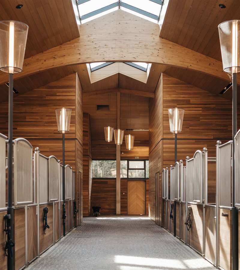 Ein heller Innenraum einer Viebrock Reithallen, der mit Holzverkleidung und großen Fenstern gestaltet ist. Der gewölbte Decke sorgt für ein offenes Gefühl, während elegante Lampen und geräumige Pferdeställe die hochwertige Bauweise unterstreichen.