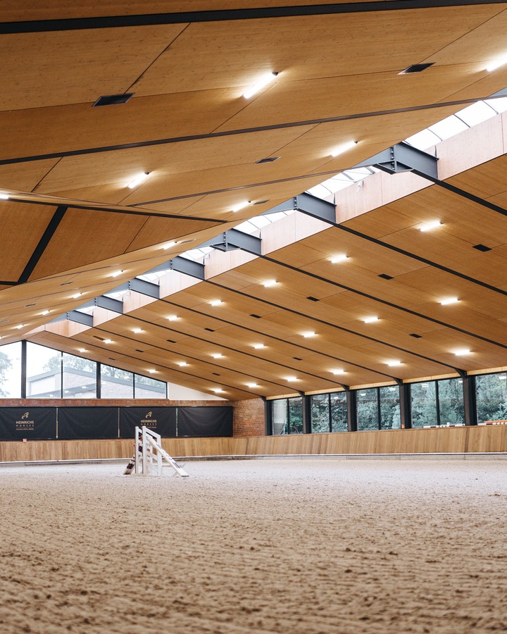 Eine moderne Viebrock Reithallen Reitbahn mit einer hellen, holzverkleideten Decke und großen Fenstern, die Licht hereinlassen. Der Boden ist sandfarben und die Wände sind mit Holzverkleidung ausgestattet. Die Arena ist mit Sprunghindernissen für das Training ausgestattet.
