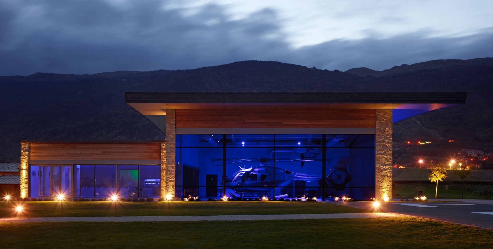 Ein modernes Viebrock Reithallen-Gebäude mit großen Glasfronten, Holz- und Steinelementen, beleuchtet mit blauen Lichtern. Im Inneren ist ein Hubschrauber sichtbar. Die umliegende Landschaft wird von sanften Hügeln geprägt, während der Abendhimmel bewölkt ist.