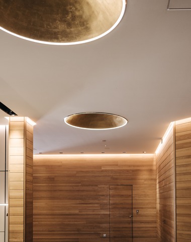 Moderne Innenansicht eines Viebrock Reithallen Gebäudes mit warmen Holzvertäfelungen und stilvollen, runden Deckenleuchten. Sanfte LED-Beleuchtung betont die Architektur. Die harmonische Gestaltung schafft eine einladende Atmosphäre.